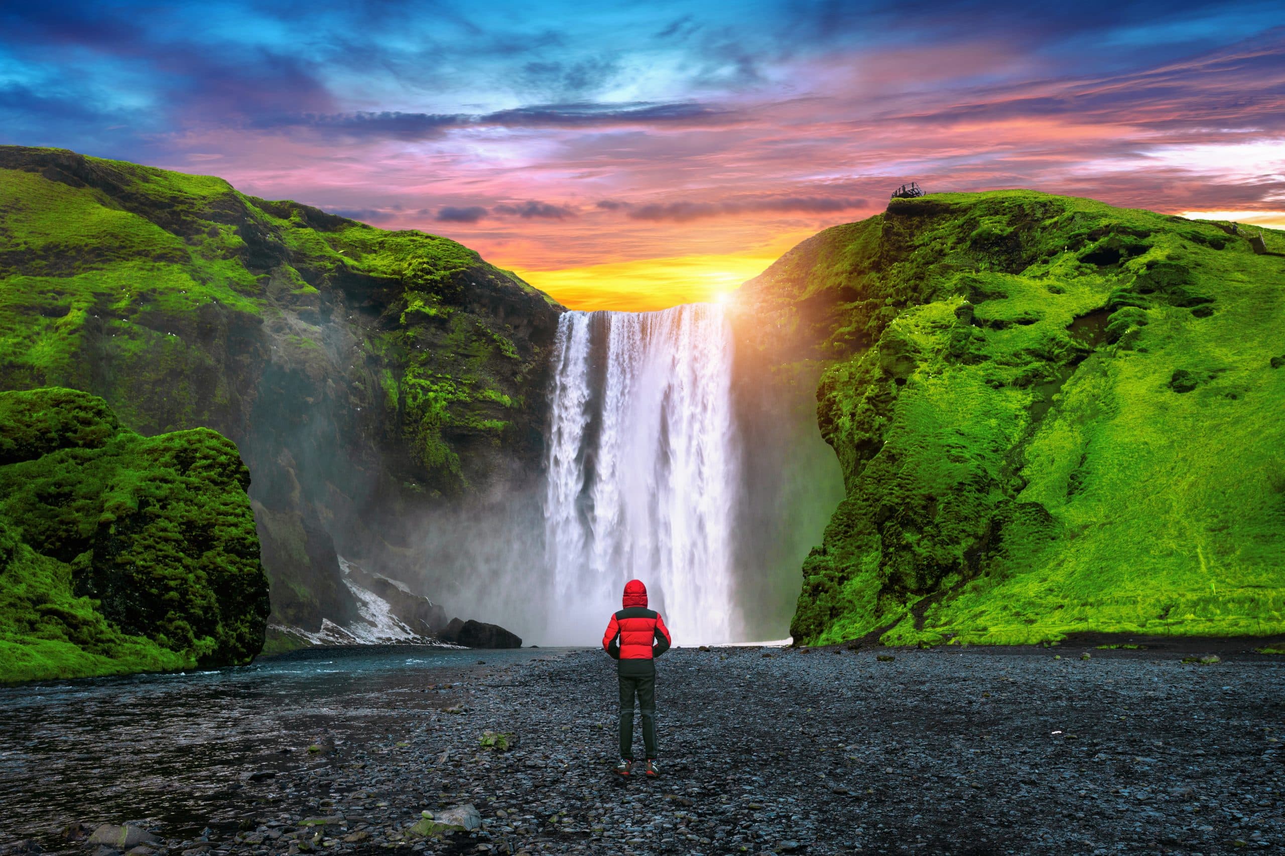 Een foto van de Skógafoss waterval in IJsland, met een reiziger op de voorgrond en een kleurrijke lucht
